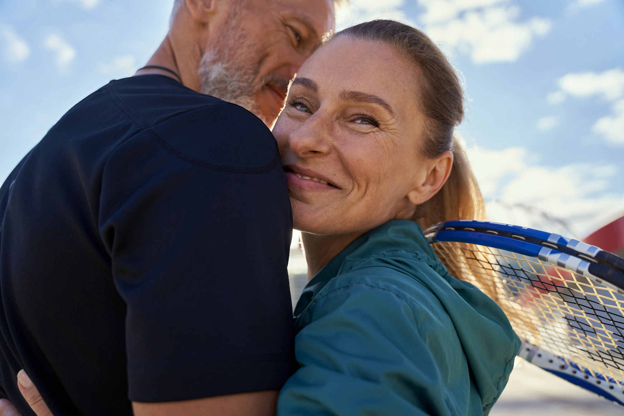 Portrait of active mature couple looking happy while embracing each other outdoors, ready for morning workout on tennis court after adrenal fatigue treatment.