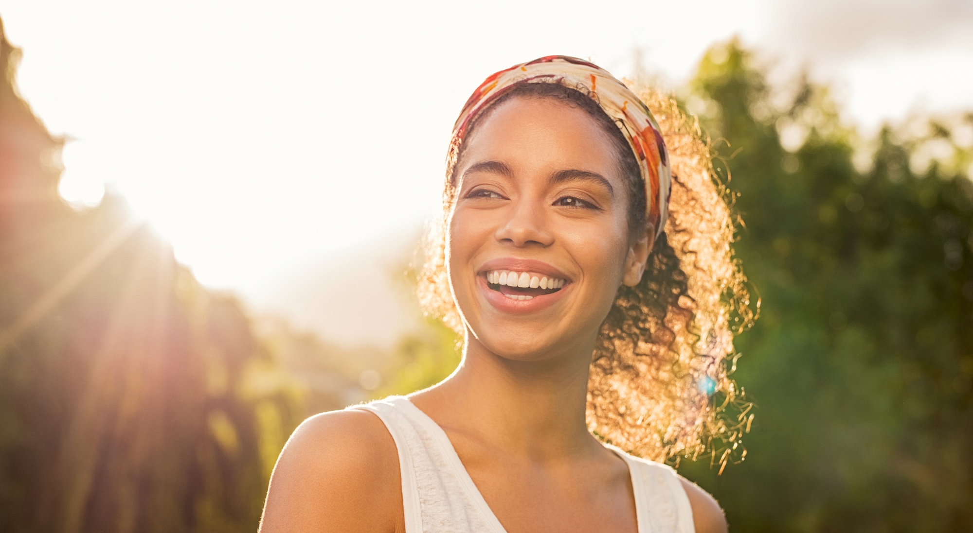 Portrait of beautiful african american woman smiling and looking away at park during sunset. Outdoor portrait of a smiling black girl. Happy cheerful girl laughing at park with colored hair band.