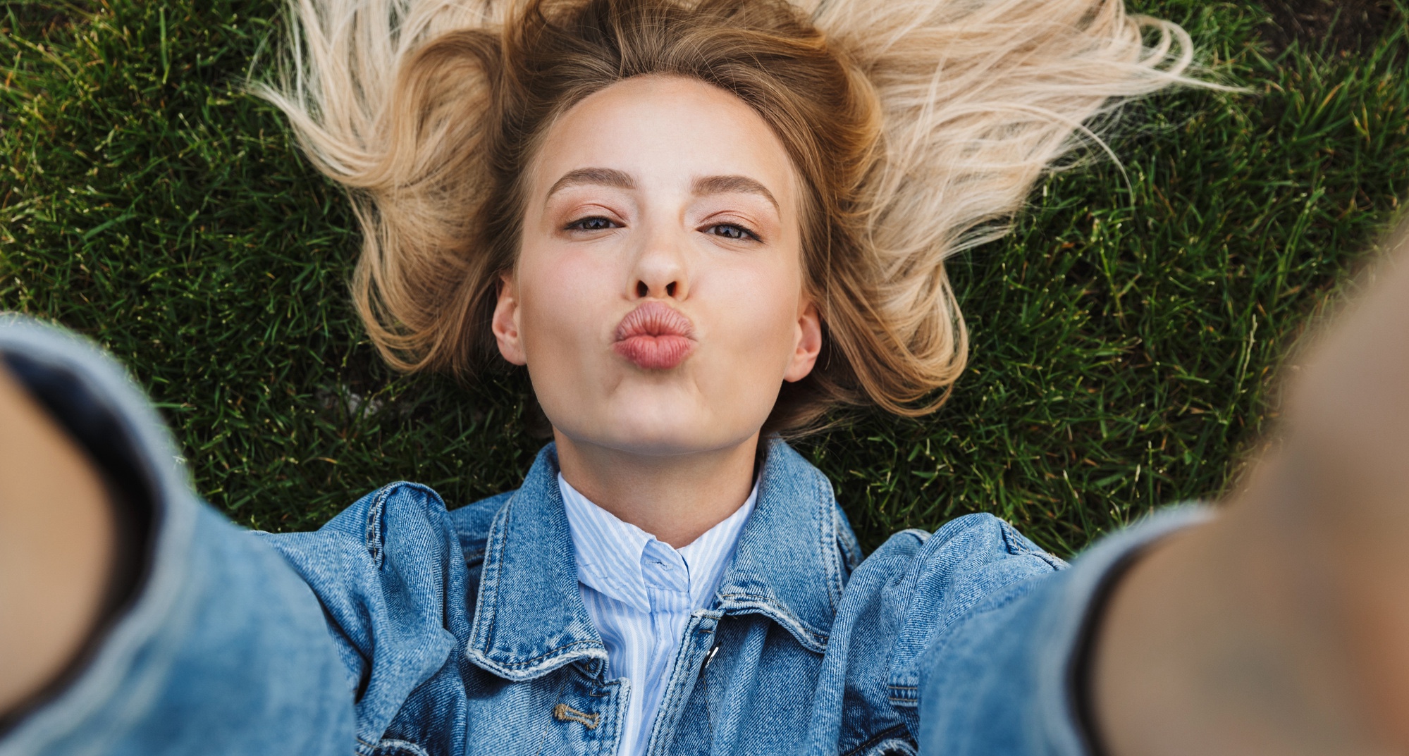 Photo of happy kind woman wearing denim jacket taking selfie photo and sending kiss while lying green grass in park