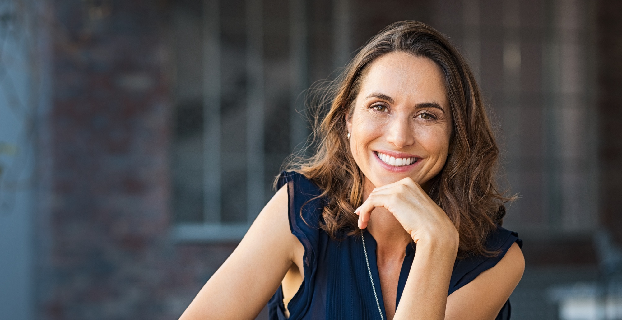 Portrait of beautiful mature woman sitting at coffee shop. Happy hispanic smiling woman sitting on a bench in outdoor cafeteria looking at camera. Portrait of carefree woman relaxing on bench.
